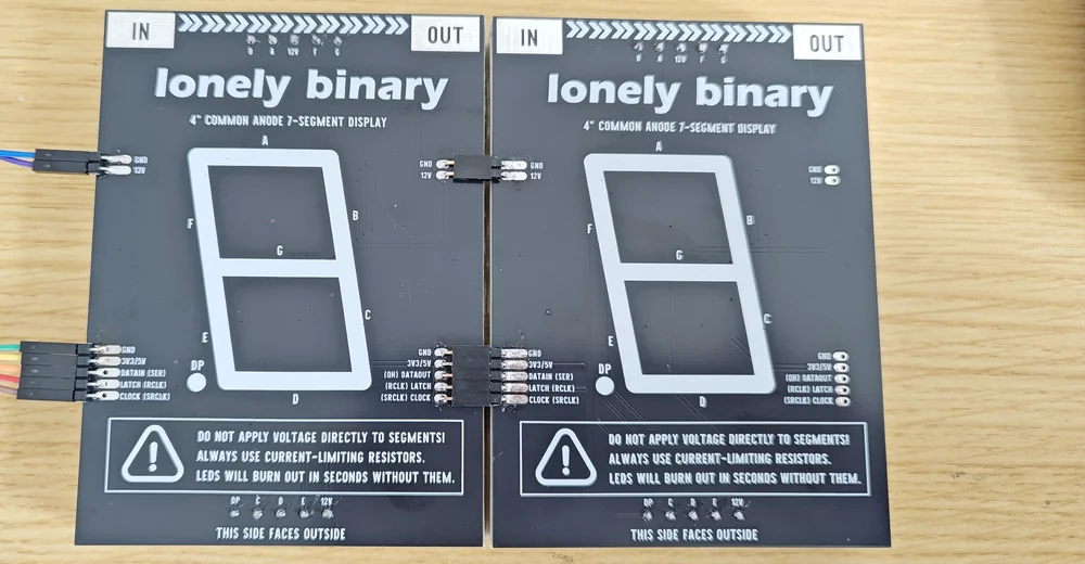 Two display modules laid back-side-up and touching edge to edge, with short black header blocks bridging the five-pin signal rows and the two-pin 12 V rows across the seam, and a ribbon of jumper wires entering the left-hand module's IN edge.