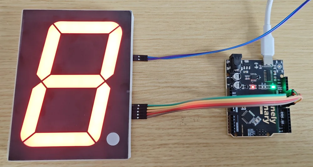 One 4-inch display module lit up showing a figure 8 in red on a dark red panel, with two ribbons of jumper wire leaving its right-hand edge and running to a black Arduino Uno compatible board at the right of the frame.
