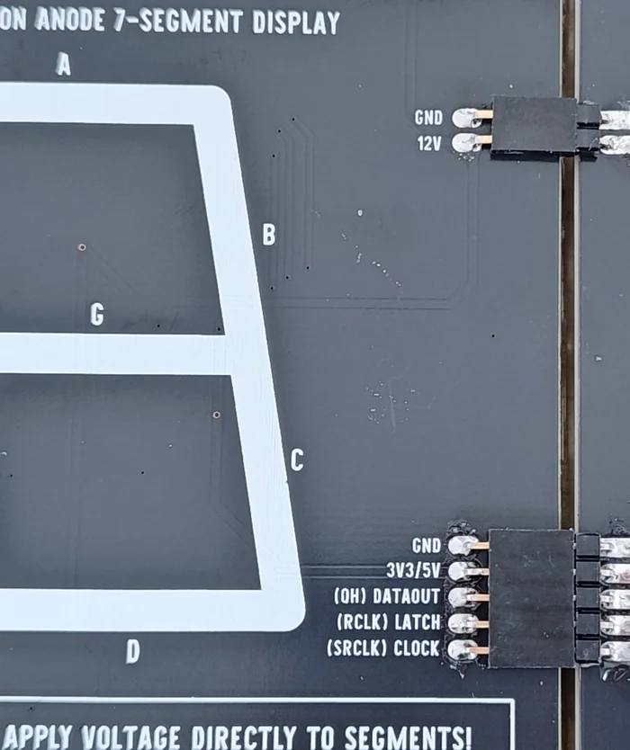 A close-up of the seam between two chained modules, back side up: the five-pin signal row of one board soldered with bright joints beside the labels GND, 3V3 5V, QH DATAOUT, RCLK LATCH and SRCLK CLOCK, a black header spacer bridging the gap, and the two-pin GND and 12V row doing the same below.
