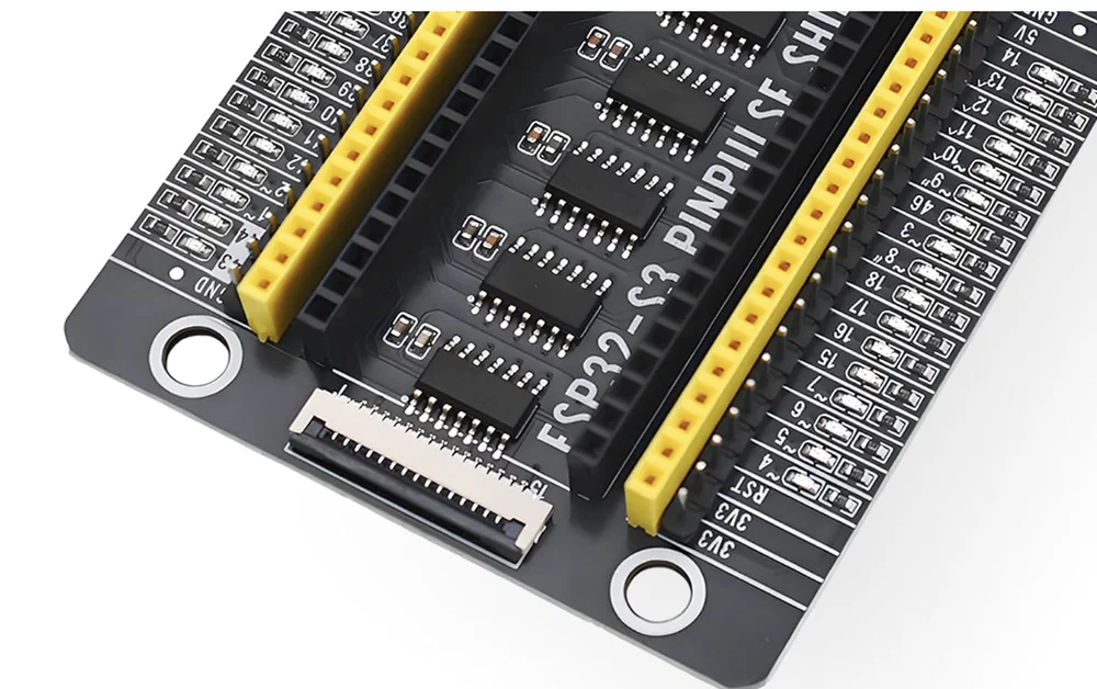 A close-up photograph of the ESP32-S3 PinPulse shield's lower half: a white fifteen-way ribbon connector at the front edge, a black board with small SOIC chips in a column, a tall black socket and a yellow female header on each side, male pins beside them, and down the right edge the labels 3V3, 3V3, RST, 4, 5, 6, 7, 15, 16, 17 and 18 each with a tiny LED.