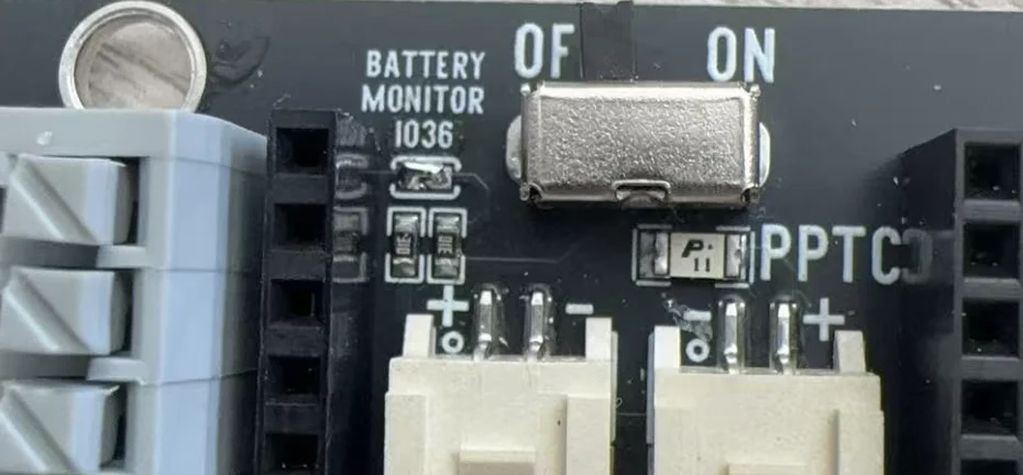 Close-up of a shield: BATTERY MONITOR IO36 printed above a small pad that has been bridged with a blob of solder, two tiny resistors side by side below it, the OFF/ON slide switch to the right, a PPTC fuse beside it, and the tops of the two white battery sockets with + and − marks.