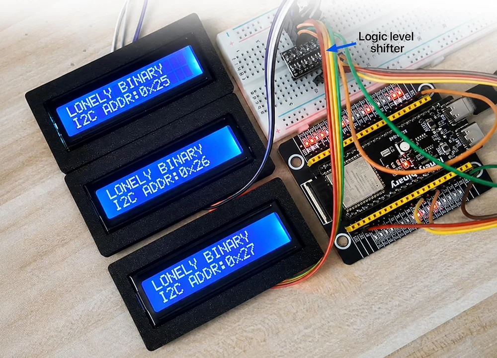 Three bezelled 1602 displays on a bench, each showing LONELY BINARY on the top row and I2C ADDR 0x25, 0x26 and 0x27 on the bottom row, all wired through one small logic level shifter on a breadboard to a single ESP32 board.