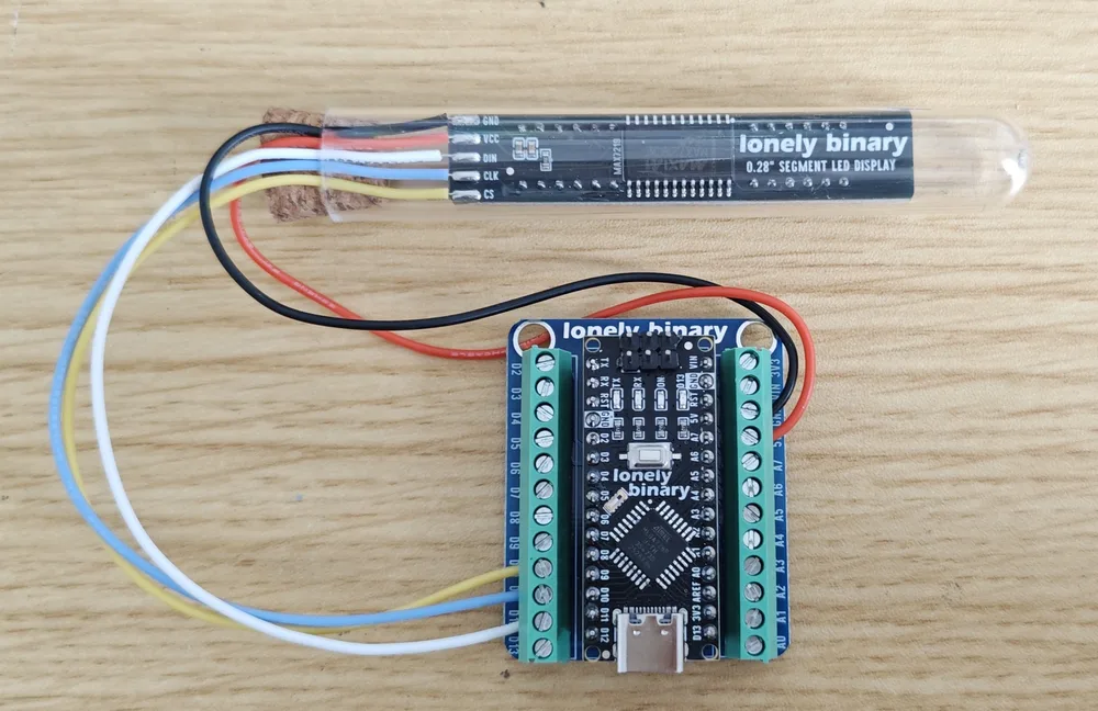 A finished eight-digit display inside a glass tube with a cork, lying on a wooden desk beside a Lonely Binary Nano expansion board with green screw terminals. Five leads run from the display's GND, VCC, DIN, CLK and CS pads to the terminal block: black to ground, red to 5 V, and white, yellow and blue for the three signal lines.