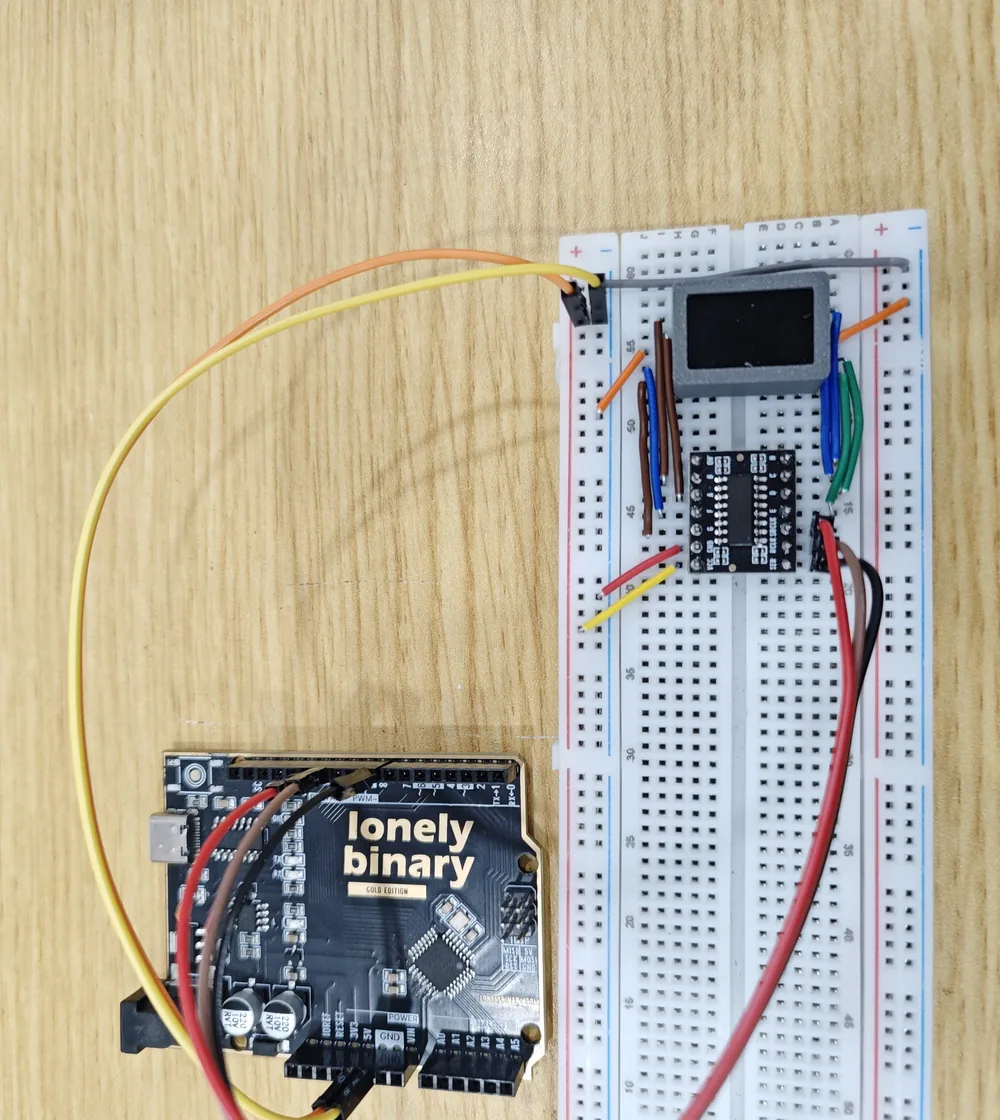 An Arduino Uno on a bench connected to a breadboard. The 74HC595 driver board sits across the breadboard's centre channel with its chip facing up, eight short jumpers run from its top and bottom rows up to a seven-segment digit in a grey 3D-printed enclosure at the top of the board, and red, brown, yellow and orange wires run from the Uno's power and digital pins.