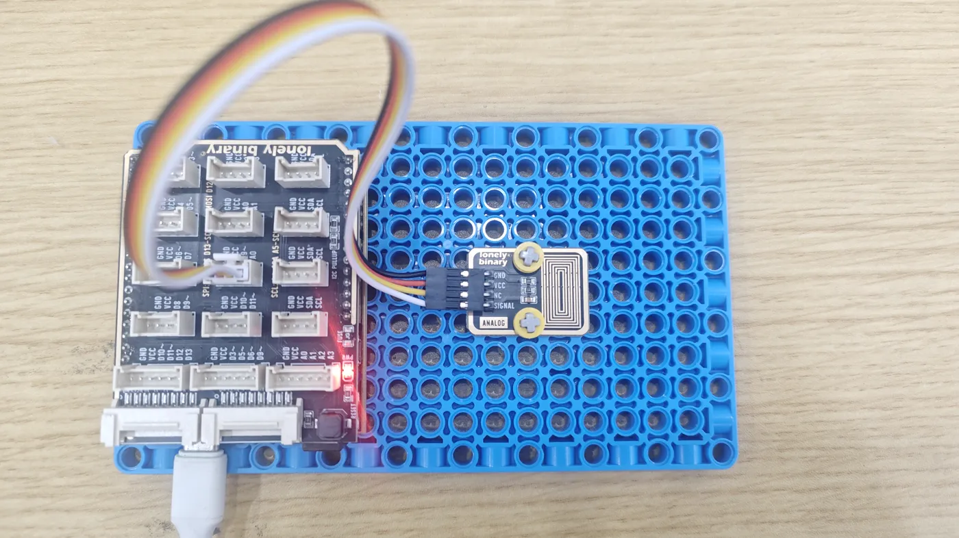 The TK65 pinned to a blue LEGO-compatible baseplate beside an Uno-format TinkerBlock shield, joined to one of the shield's four-pin sockets by a four-wire cable into its GND, VCC, NC and SIGNAL pins, the gold loops facing up.