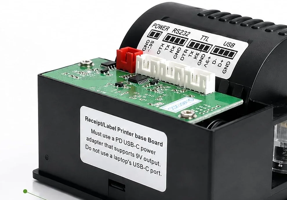 The label printer module from behind: a black square case with a driver board across the top, a printed legend naming four connector groups — POWER with GND and 9V, RS232 with DTR TX RX GND, TTL with DTR TX RX GND, and USB with GND, D plus, D minus and 5V — and a white sticker on the case warning that a PD USB-C adapter supporting 9V output must be used and not a laptop's USB-C port.