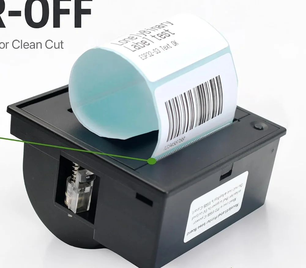 A printed label curling out of the top of the label printer: a white label on a pale blue backing strip carrying the words Label test, ESP32-S3 Text OK and a barcode, with a close-up inset showing the serrated tear bar the label is pulled across.